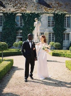 a bride and groom walking in front of a large building with hedges on both sides