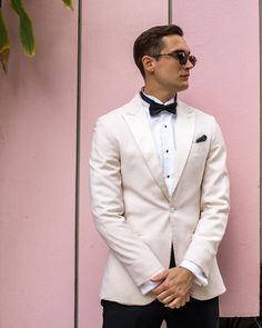 a man in a white tuxedo stands against a pink wall