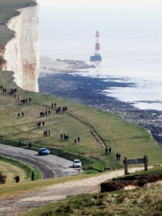 people are walking on the side of a road near the ocean and a light house