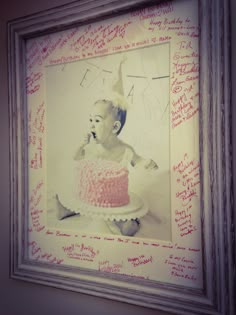 a framed photograph of a baby holding a cake with pink frosting and writing on it