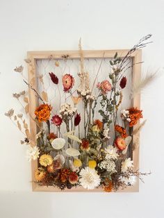 an arrangement of flowers in a wooden frame on a white wall with dried grass and leaves