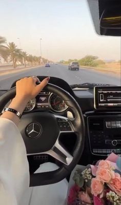 a woman driving a car on the road with flowers in front of her and palm trees behind her