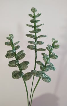 two green plants are in a vase on a table with white wall behind them and one is made out of wire