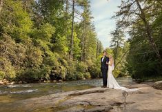 a bride and groom standing on rocks in the middle of a river surrounded by trees