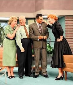 four people are standing together in front of a tv set, one is wearing a suit and the other wears a dress