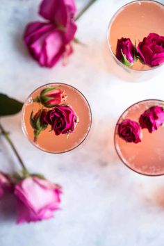 three glasses filled with pink flowers on top of a white countertop next to purple roses