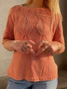 a woman in an orange top making a heart shape with her hands while standing next to a wall