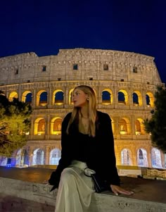 a woman sitting on a ledge in front of the colossion at night time