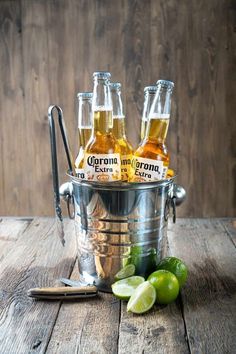 four bottles of corona beer in an ice bucket with limes and a spoon on a wooden table