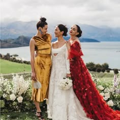 three women standing next to each other in front of some flowers and water with mountains in the background