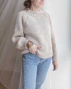 a woman standing in front of a window wearing a white sweater and jeans with her hand on her hip