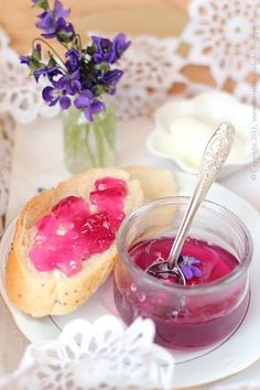 a jar of jelly next to a plate with bread and purple flowers in the background