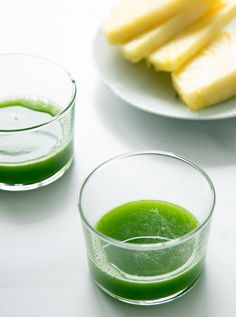 two glasses filled with green liquid next to slices of pineapple