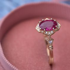 a close up of a ring in a pink box with diamonds on the sides and an oval shaped ruby stone surrounded by small white stones