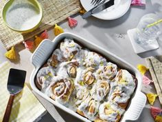 a white dish filled with cinnamon rolls on top of a table next to plates and utensils