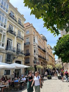 people are walking down the street in front of some buildings and tables with umbrellas