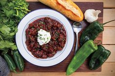 a white plate topped with chili next to green peppers and bread on top of a wooden table