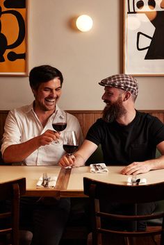 two men sitting at a table with wine glasses in their hands and one man smiling