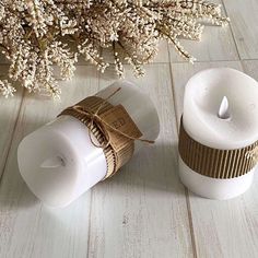 two white candles sitting next to each other on a wooden table with dried flowers in the background