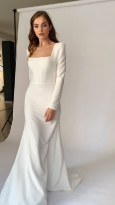 a woman standing in front of a white backdrop wearing a long sleeved wedding dress