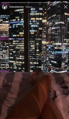 a person laying in bed with the city lights lit up behind them at night time