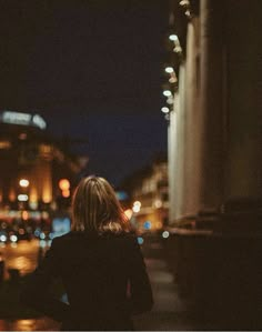 a woman is standing on the sidewalk at night looking out to the street lights in the distance