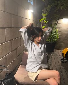 a woman sitting on top of a wooden bench next to a potted plant in front of a brick wall