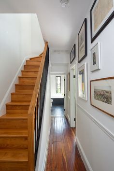 a hallway with wooden stairs and pictures on the wall next to wood flooring in front of white walls