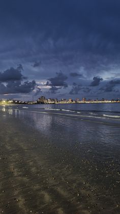 the sky is very dark and cloudy over the water at night on the beach with buildings in the distance