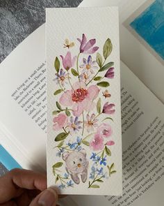 a hand holding an open book with watercolor flowers on it and a bear in the middle