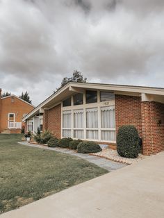 the front of a brick house with grass and bushes in front of it on a cloudy day