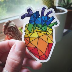 a hand holding a sticker with a colorful heart on it in front of a potted plant