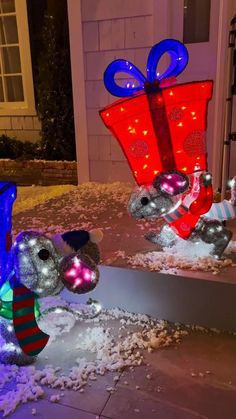 two christmas decorations in front of a house with snow on the ground and presents wrapped in lights