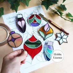 a hand holding a piece of paper with stained glass ornaments on it next to scissors and other crafting supplies