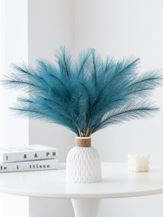 a white table topped with a vase filled with blue feathers next to two books and a candle