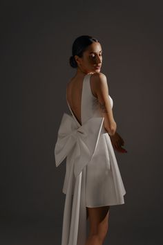 a woman wearing a white dress with a large bow on the back, standing in front of a gray background