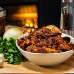 pulled pork in a white bowl next to garlic and parsley on a cutting board