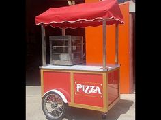 a pizza cart with a red awning on the top and wheels parked in front of an orange building