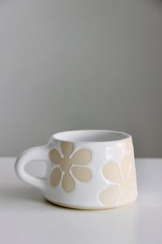 a white cup with brown flowers on it sitting on top of a table next to a wall