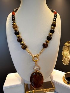 The necklace in the image showcases a striking central amber-colored oval pendant, suspended from a gold-tone ring that also holds a smaller, translucent pearl, creating a dual charm effect. The beads, in varying shades from deep browns to warm amber and occasional hints of red, are faceted for a gentle sparkle. The assembly is completed with a gold-tone beads for a touch of elegance followed  by tiger eye  12 mm grade A beads and 8mm of black obsidian , gold faceted rondelles, red agate represe Luxury Amber Oval Necklace, Elegant Amber Pendant Necklace, Elegant Oval Amber Necklaces, Elegant Brown Pendant Jewelry, Gold Teardrop Pendant Necklace With Gemstone Accents, Elegant Amber Round Pendant Jewelry, Luxury Amber Jewelry With Natural Stones, Gold Oval Necklace With Gemstone Accents, Gold Necklace With Oval Gemstone Accents