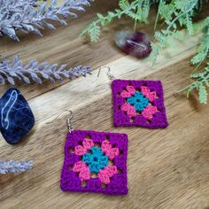 two square shaped crocheted earrings sitting on top of a wooden table next to plants