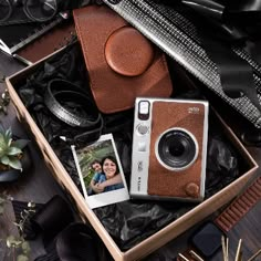 an open box containing a camera, photos and other items on a wooden table next to plants