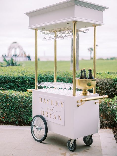 A white wedding bar cart with gold accents, featuring a champagne holder, glass display, and a personalized sign. Outdoor Wedding Bar Ideas, Bourbon Bar Wedding, Bar Setup Ideas, Wedding Bar Setup, Elegant Bar Cart, Wedding Bar Cart, White Bar Cart, Bar Ideas Wedding, Champagne Holder