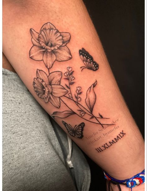 Black and white tattoo of two daffodils intertwined with smaller flowers on the forearm. There is a small butterfly to the right of the flowers, and Roman numerals underneath.  Everything has light grey shading and white highlights. Daffodil Half Sleeve Tattoo, Narcissus Flower Tattoo With Butterfly, Tattoo With Lily Flowers, Flower Tattoo With Roman Numerals, Butterfly Daffodil Tattoo, Flower And Roman Numeral Tattoo, Daffodil Name Tattoo, Daffodil With Butterfly Tattoo, Daffodil Hand Tattoo