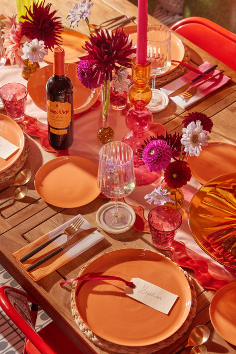 1. A table runner in shades of pink and red create the base of the tablescape. The clashing shades and tones will give the overall tablescape a playful mis-matched vibe.

2. Place a variety of candlesticks and small, multi-coloured vases of flowers dotted down the runner. 

3. Layer terracotta orange shaded plates over woven rattan placemats for an organic feel. Brassy cutlery and dip-dyed napkins bring additional pops of warmth and texture. Pink Orange Yellow Tablescape, Red Pink Orange Party Decor, Pink Orange Table Setting, Colorful Place Setting, Colorful Table Runner, Modern Mexican Table Setting, Pink Tablescape Party, Pink And Orange Dinner Party, Pink And Orange Tablescape