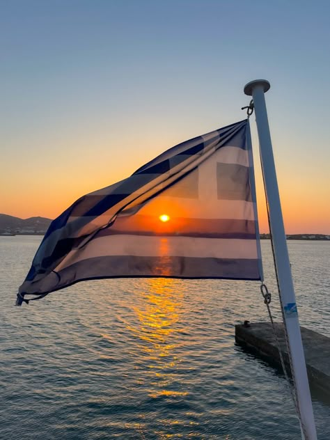 Multiple colors of bright orange red and yellow sunset of the ocean of Greece with the Greek flag shaded over flying in the wind Greece Flag Aesthetic, Greek Flag Aesthetic, Visonboard 2025, Greek Sunset, Ef Ultimate Break, Greece Aesthetics, Greece Sunset, Aesthetic Greece, Greek Aesthetic