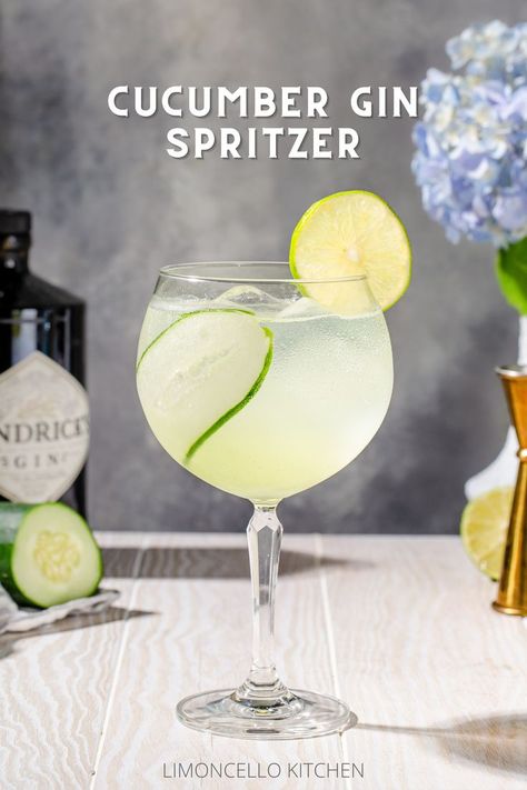 Side view of a Cucumber Gin Spritzer cocktail on a white wood countertop. The drink is in a tall stemmed glass with a round bowl. It is light green and semi translucent, and the glass has a strip of cucumber inside and a lime wheel on the rim. In the background are bar tools and hydrangea flowers along with ingredients. Fancy Drink Garnish, Cucumber Spritzer Vodka, Cucumber Drink Alcohol, Cucumber Spritzer, Gin Cocktails Summer, Light Summer Cocktails, Cucumber Gin Cocktail, Strawberry Mojitos, Grinch Recipes