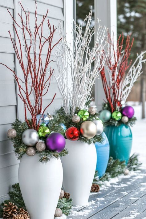 White and blue vases feature festive arrangements of colorful ornaments, pine branches, and red and white twigs, set on a snowy wooden porch. Christmas Porch Planters Diy, Outdoor Xmas Planter Ideas, Cheap Christmas Porch Decor, Diy Christmas Pots Outside, Diy Christmas Planter Ideas, Outdoor Christmas Planters Winter Porch Front Doors, Winter Wonderland Outside Decorations, Gnome Christmas Decor Ideas, Christmas Garden Decorations Outdoor