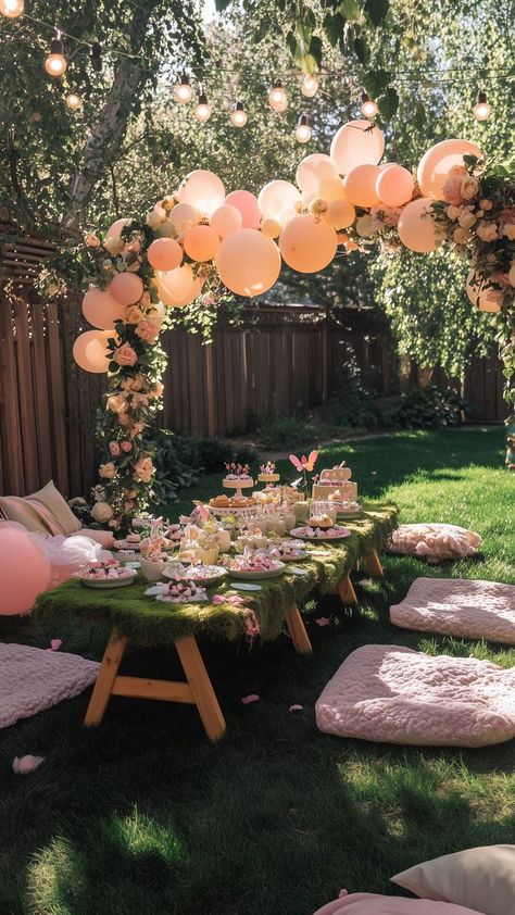 Outdoor fairy garden birthday party setup with a moss-covered table, soft pink cushions, whimsical balloon arch, string lights, and delicate floral decorations, creating a magical and enchanting atmosphere. Leaf Birthday Theme, Park Tea Party Birthday, Outdoor Fairy Birthday Party Ideas, Enchanted 1st Birthday Theme, Enchanted Forest Themed Birthday Party, Secret Garden Decorations Party, Girls Garden Party Birthday, Rose Garden Birthday Theme, Fairy Garden Gender Reveal