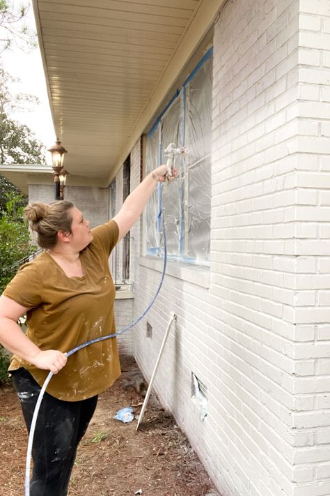 Woman using Graco Magnum 262800 X5 Stand Airless Paint Sprayer to paint exterior brick home white with paint sprayer. Instructions of how to paint a brick house with pros and cons. Blue painters tape with sheeting covering windows. Exterior Painted Brick House, Painted White Brick House, Exterior Painted Brick, Home Exterior Updates, Painting Brick Exterior, Paint House Exterior, Red Brick Paint, Brick Painted White, Brick Exterior Makeover